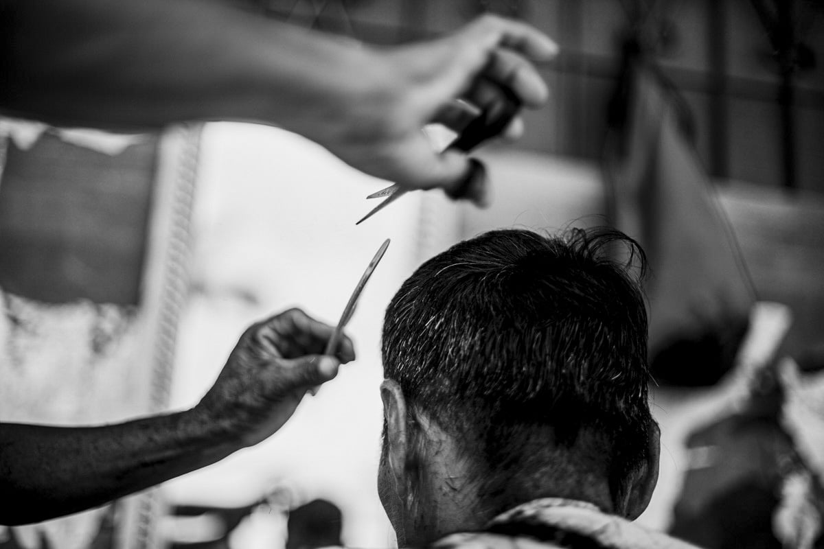 The barber’s arms reach into the frame from the left, scissors and comb poised over the client’s head. The barber’s hands, blurred with motion, contrast with the clear, still outline of the client's head. His hair, neatly shorn, covers the back of his neck.