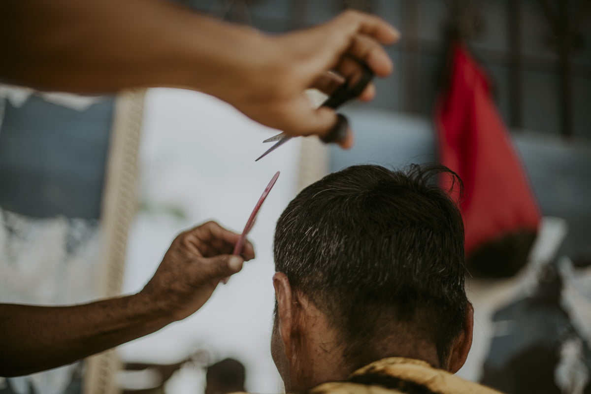 The barber’s arms reach into the frame from the left, scissors and comb poised over the client’s head. The barber’s hands, blurred with motion, contrast with the clear, still outline of the client's head. His hair, neatly shorn, covers the back of his neck.