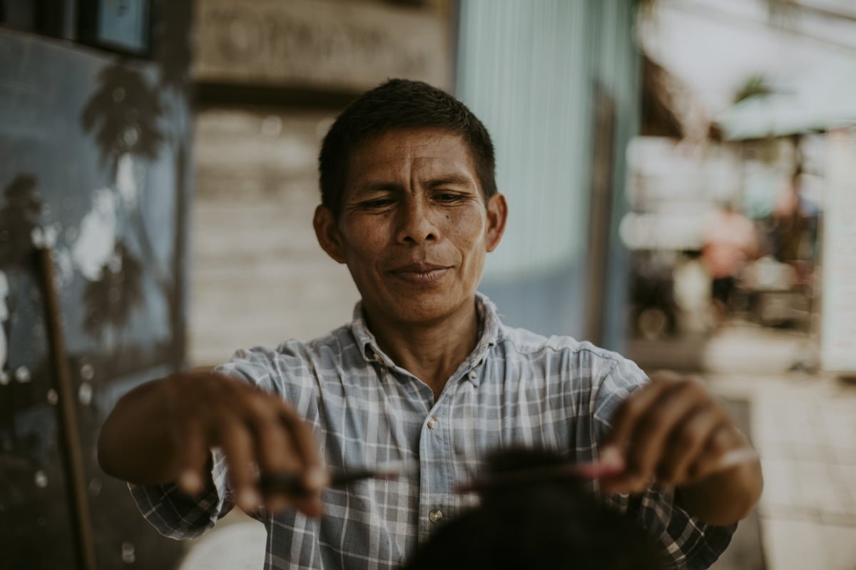 A portrait of the indigenous barber at work, this image captures his wide cheekbones, the hint of a smile, and his eyes lowered in concentration over his client. His own haircut is neat. The collar of his plaid shirt is worn.  His arms reach toward the viewer, his scissors and comb hovering over the head of the man in the chair, his face and body conveying a sense of ease. 