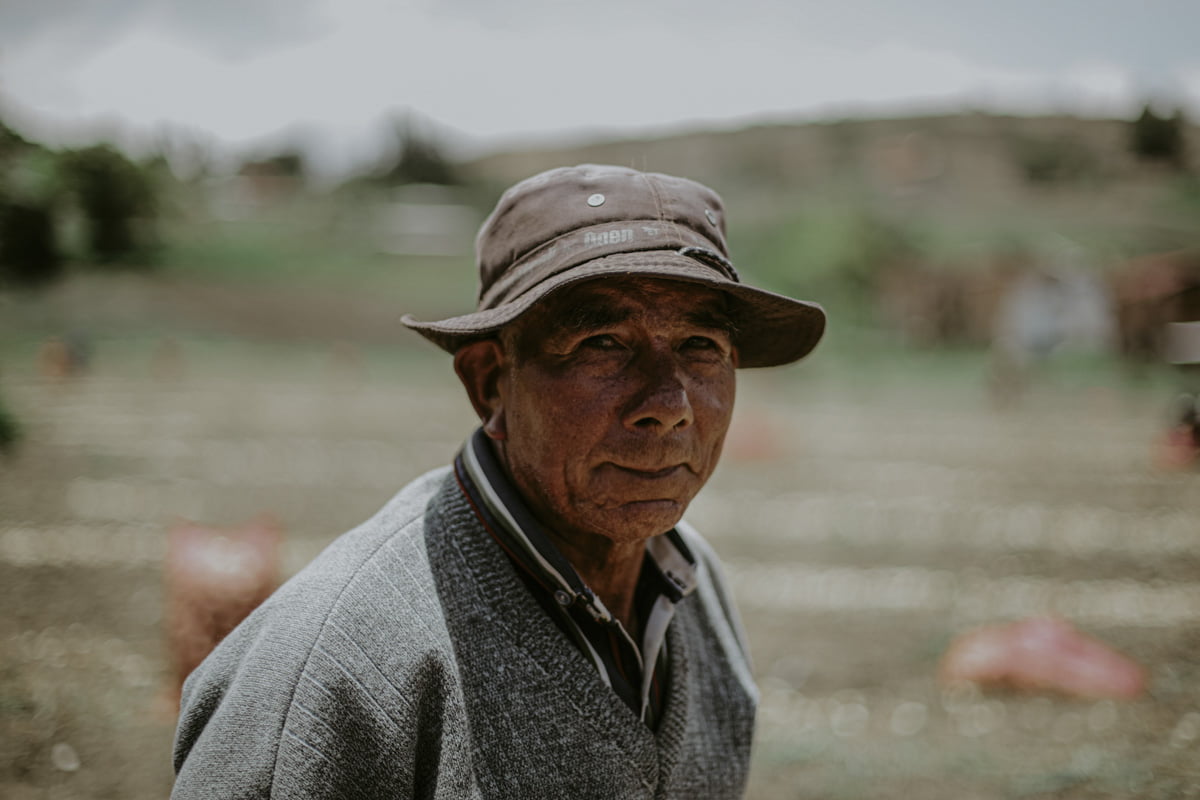 In this portrait, an indigenous, round faced onion picker is dressed in a v-neck sweater with a button down underneath, and a bucket hat that pushes down, splaying his ears. The onion field, a haze behind him, with no other workers in sight, reflects the solitary nature of this work. He looks gently, but intently at the camera. 