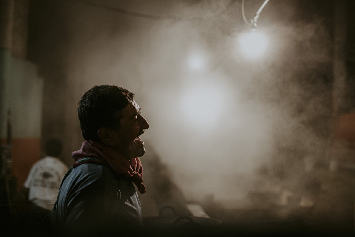 A profile of a worker in the now smoke filled factory, which is backlit by the bright glare of an overhead light. He is open mouthed,  either from speaking or laughing, a moment of respite from the heat and labor this process demands. 