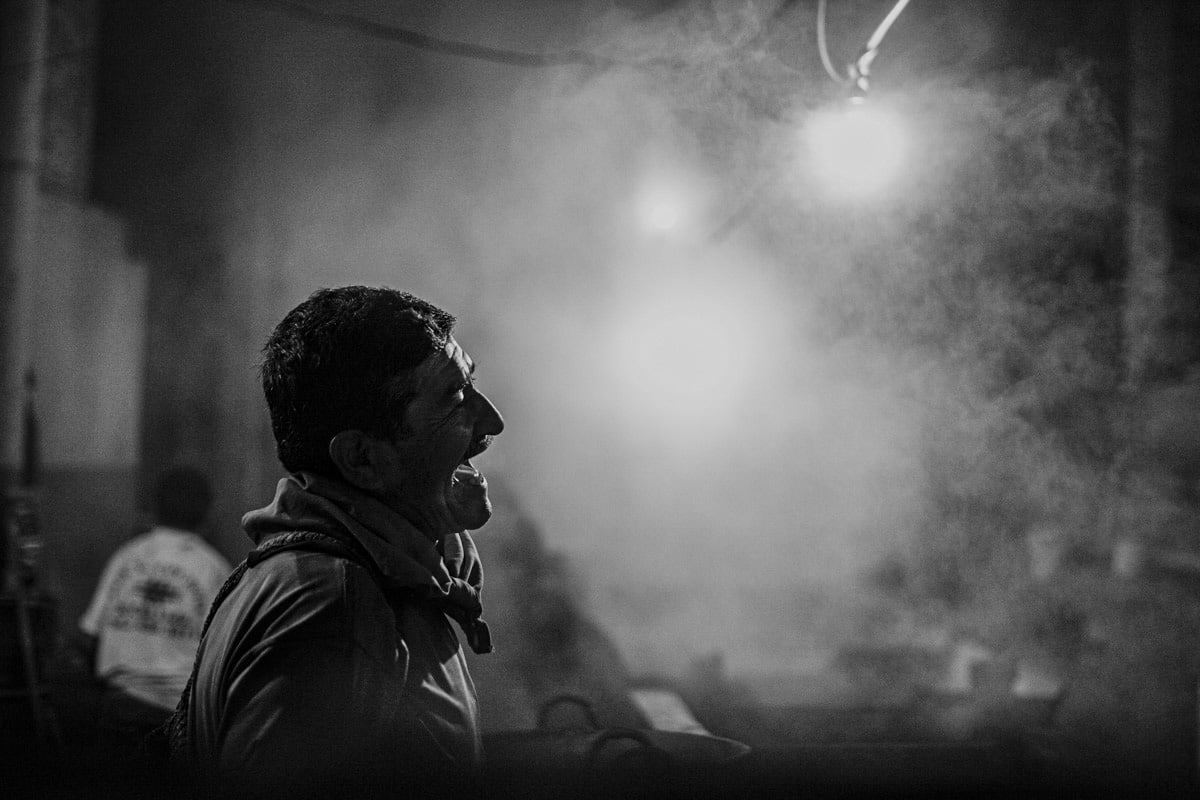 A profile of a worker in the now smoke filled factory, which is backlit by the bright glare of an overhead light. He is open mouthed,  either from speaking or laughing, a moment of respite from the heat and labor this process demands. 