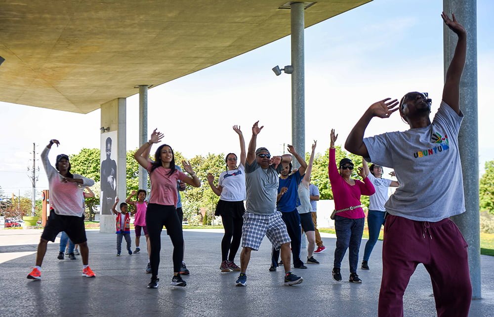 Mafa leads 15 participants in a dance class. This photo captures everyone with their arms up and walking forward.