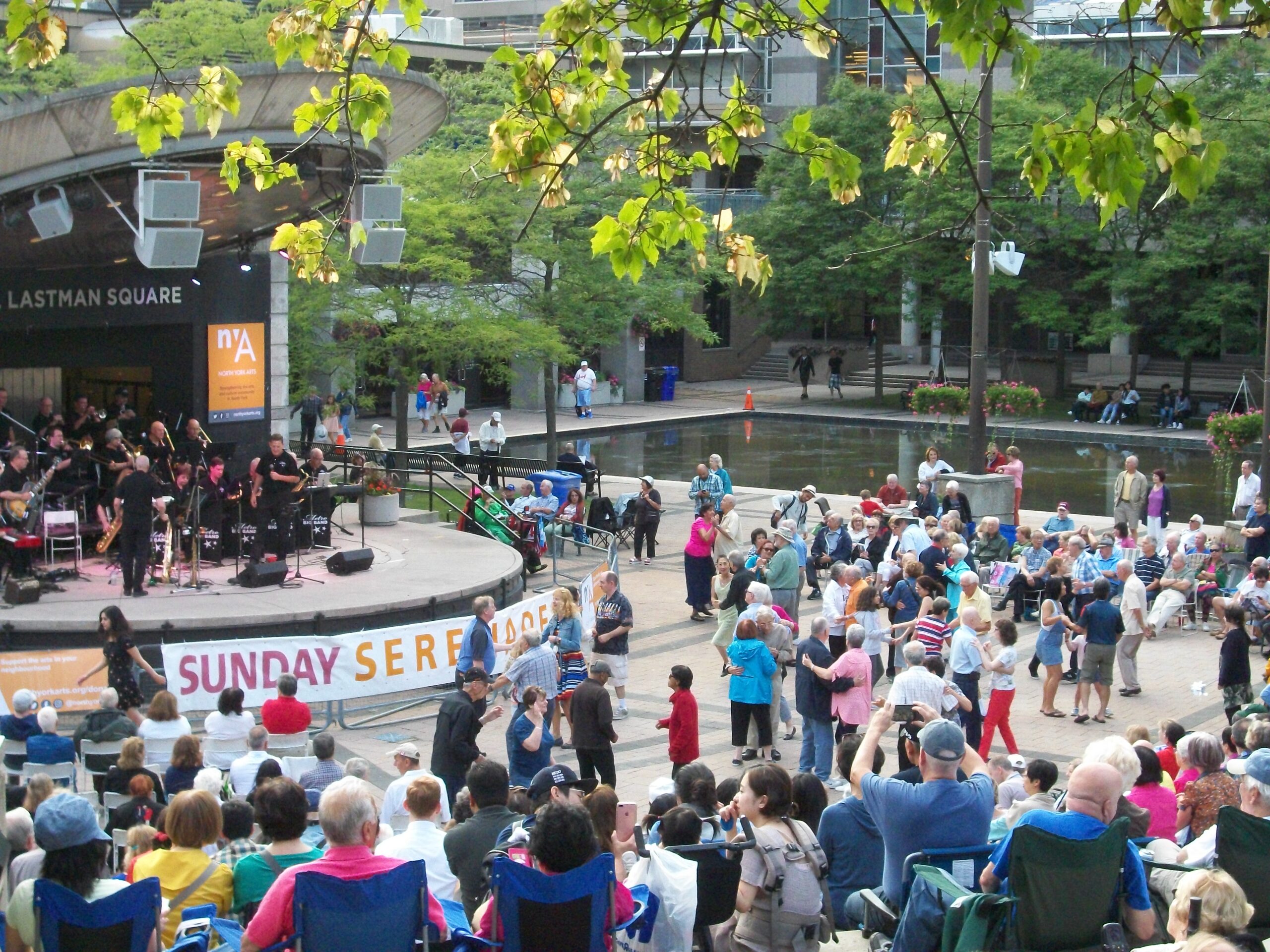 A photo of Mel Lastman Square presenting Sunday Serenades in 2018. There is music on the stage and a happy crowd.